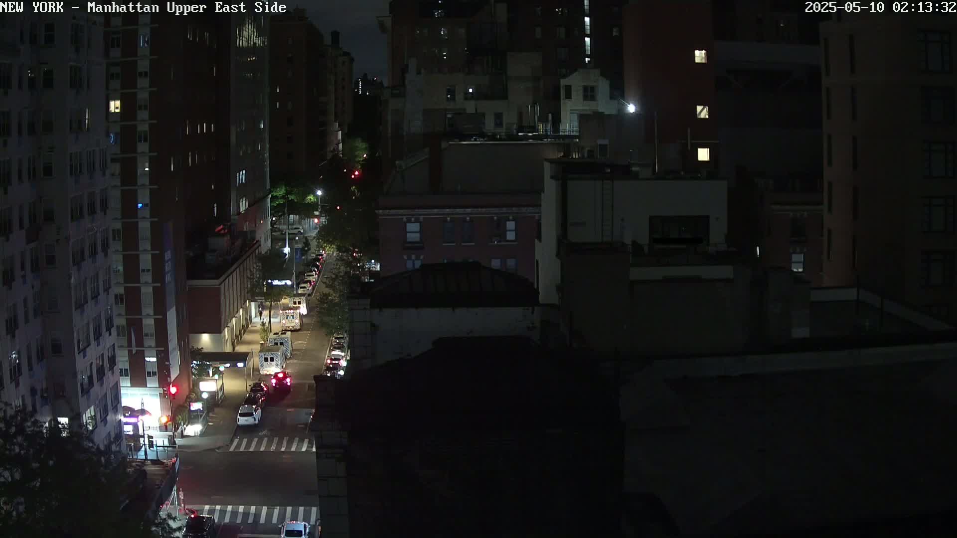 A nighttime aerial view of a city street shows several dark buildings, sparse traffic, and a few illuminated streetlights.