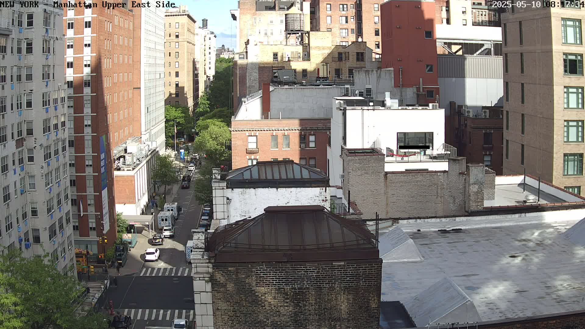 A sunny city street scene shows several multi-story brick buildings, some with visible rooftops, and cars parked along a street.