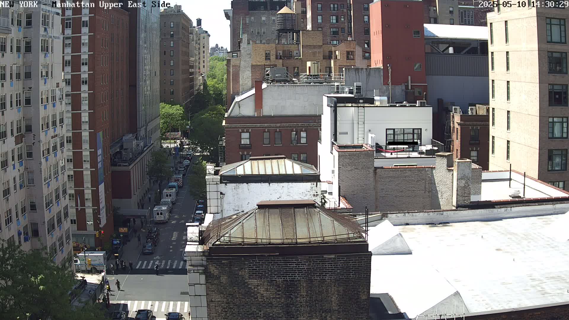 A high-angle, sunny view of a city street with tall brick buildings and parked vehicles, some of which are emergency vehicles.