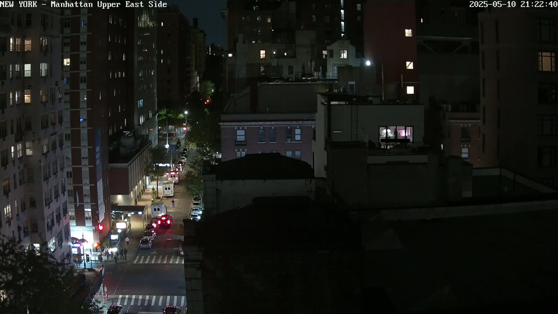 A nighttime aerial view of a city street shows several multi-story buildings with some lights on, vehicles on the street, and people walking on the sidewalk.