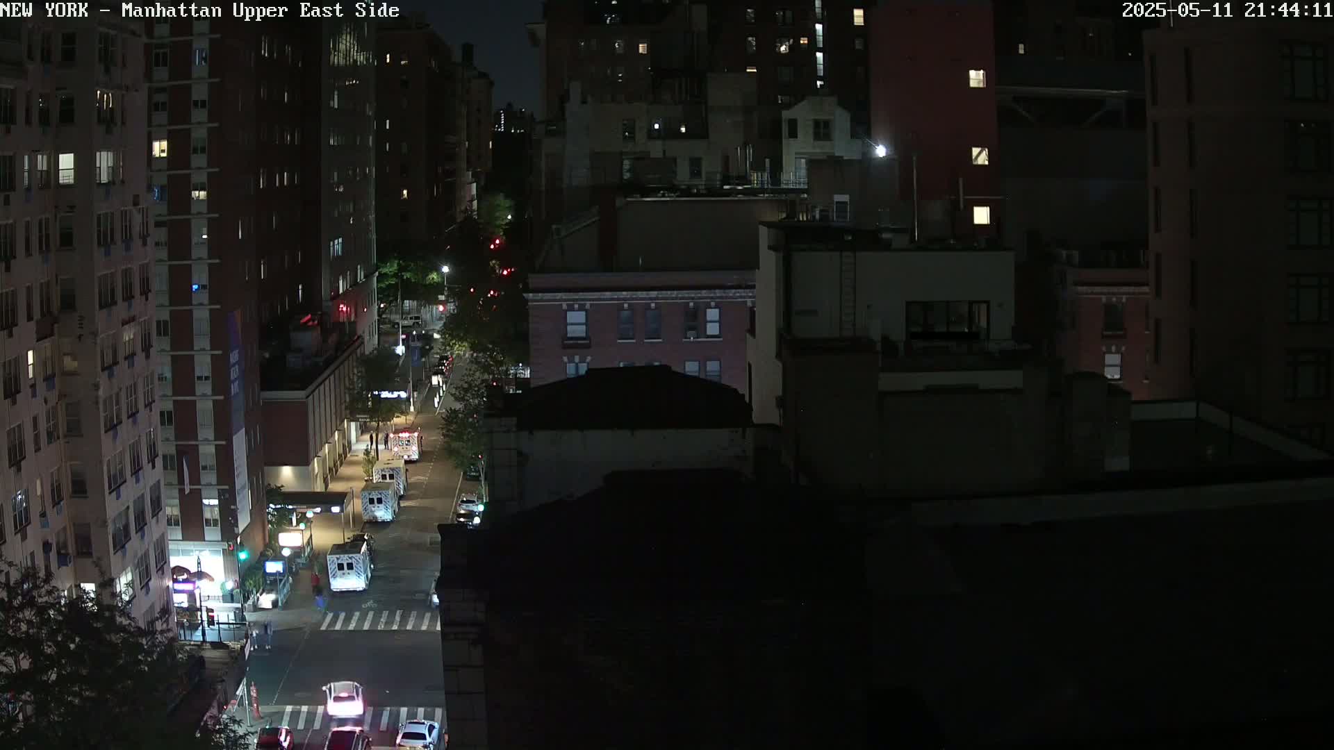 A nighttime aerial view shows city streets with several vehicles, including what appear to be emergency vehicles, parked near tall buildings under dark, clear skies.