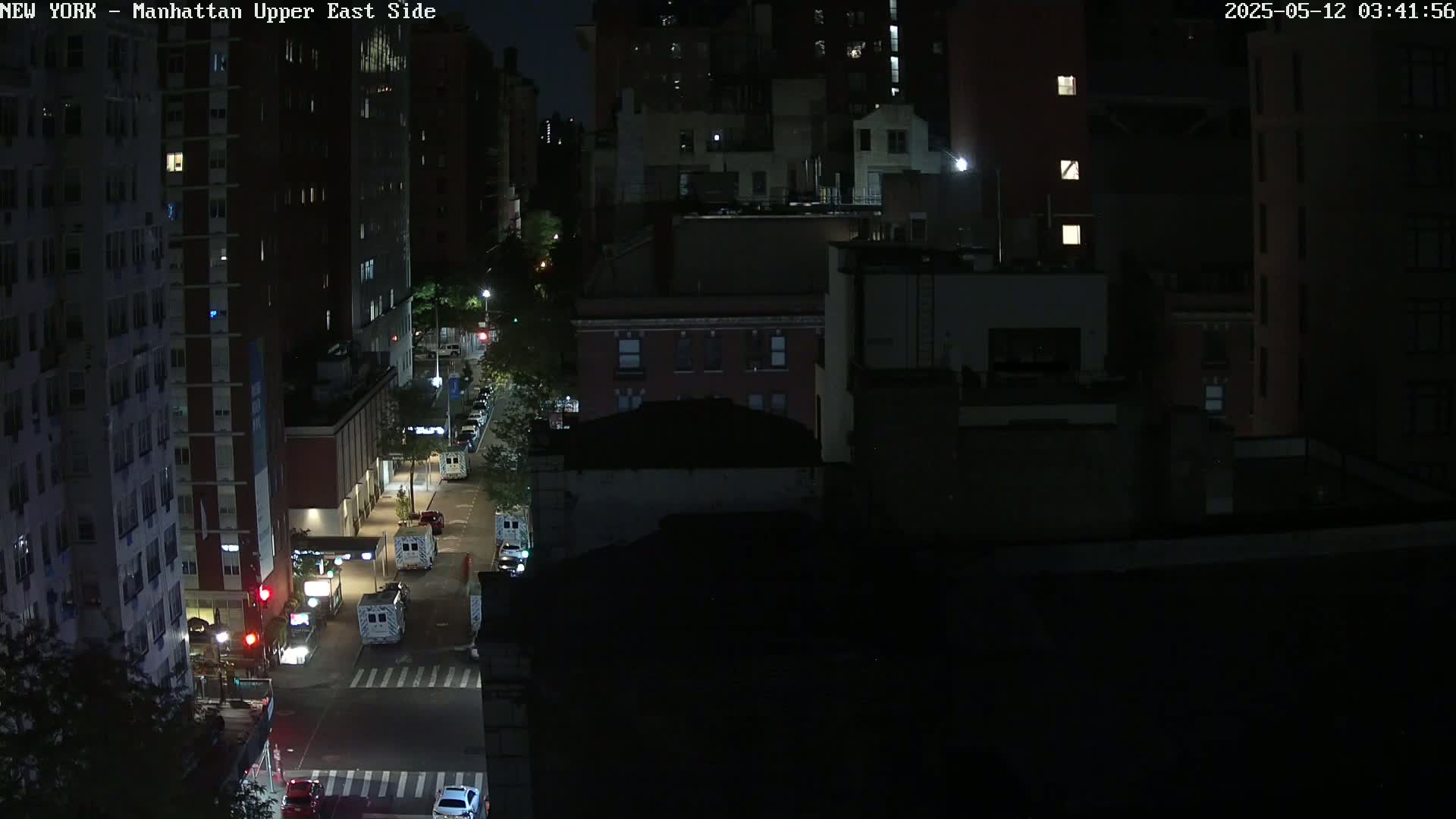A nighttime aerial view of a city street shows several vehicles parked along the side of the road near tall buildings.