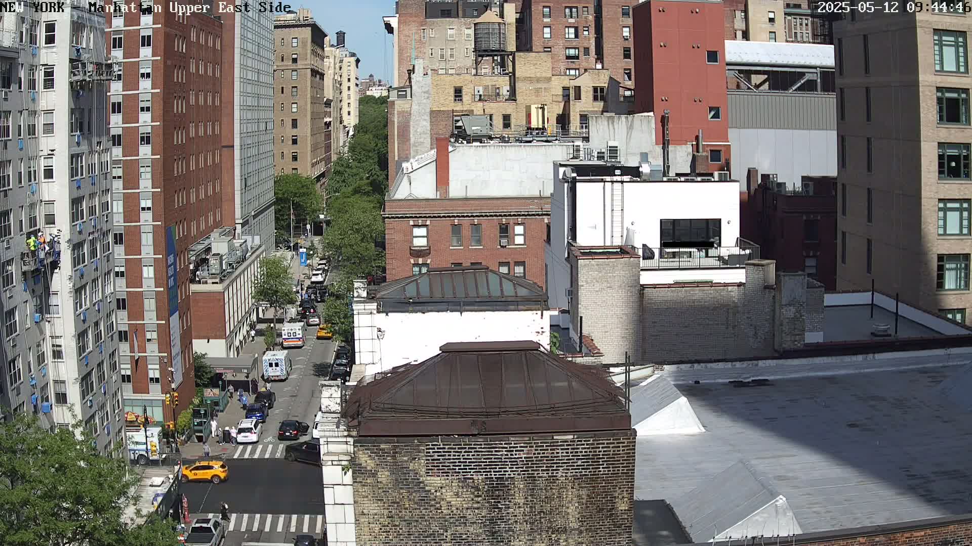 A sunny city street scene shows a high-angle view of numerous brick buildings, some under construction, with cars parked along the street and a few vehicles in motion.