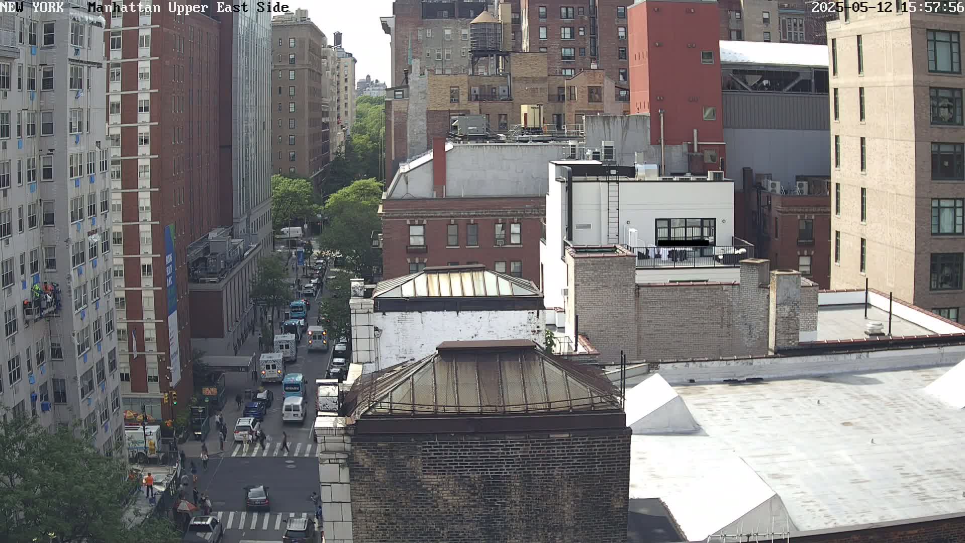 A sunny city street scene shows several multi-story brick buildings, a street with cars and pedestrians, and scaffolding on the side of one building.