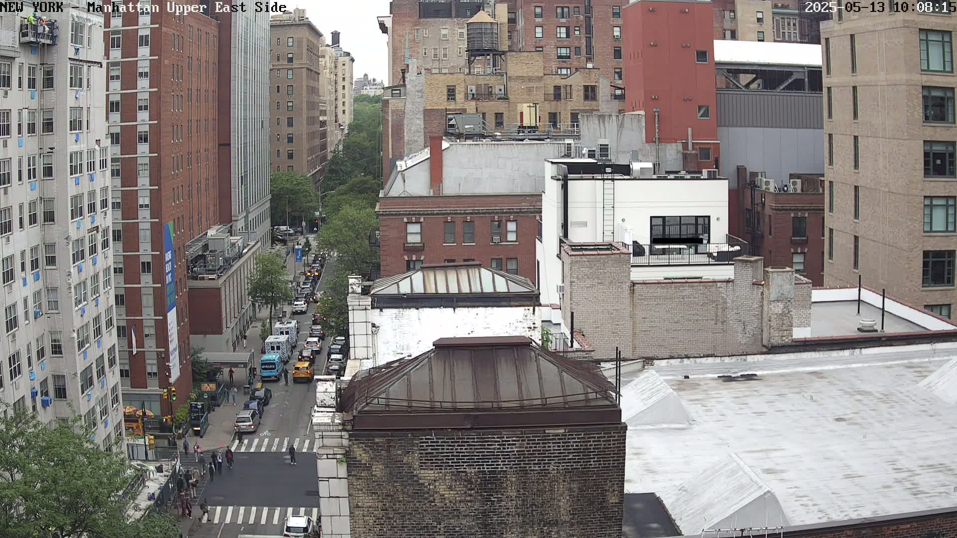 A high-angle view of a city street with numerous brick buildings on a cloudy day, showing vehicles parked and driving on the street below.