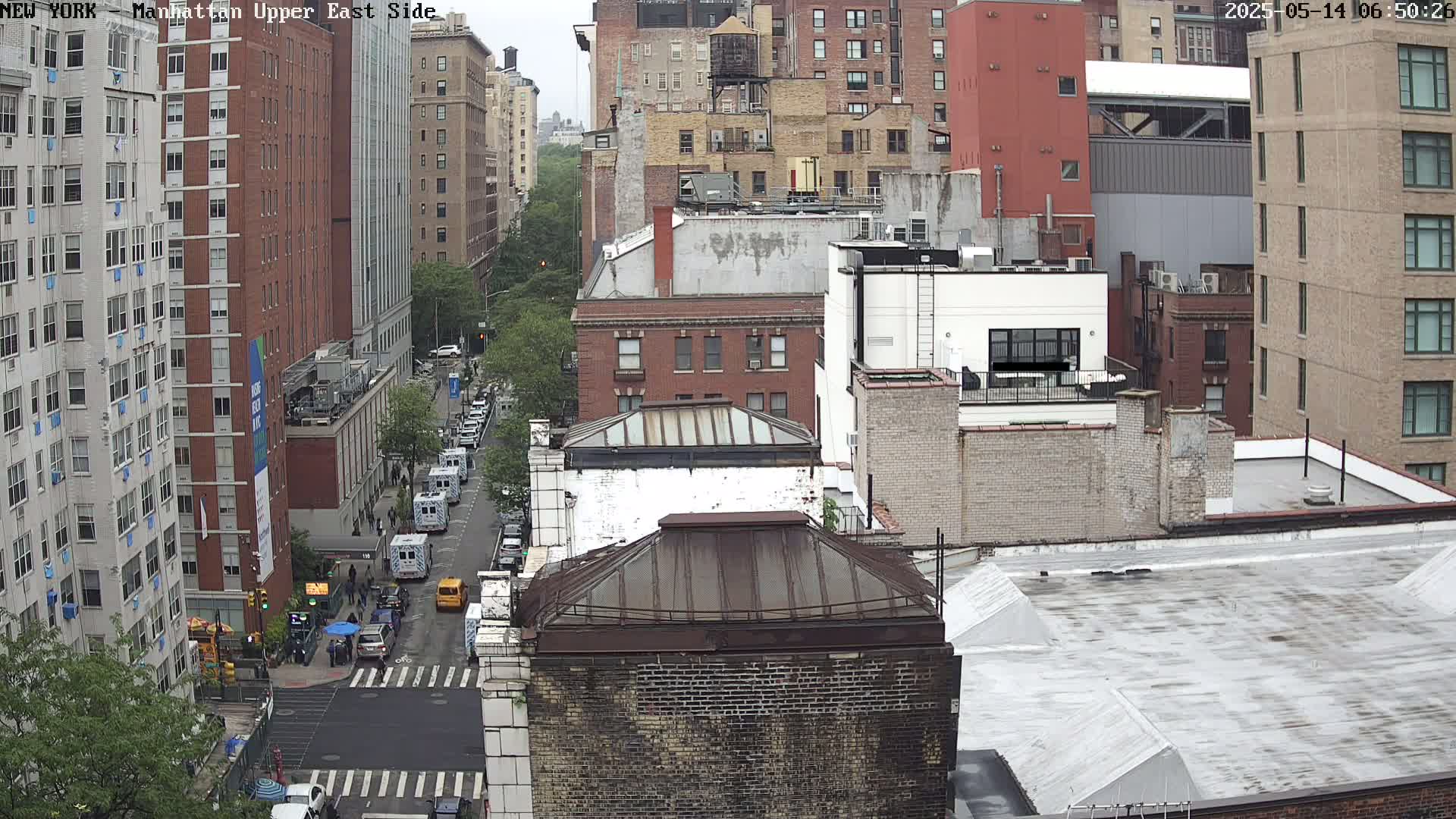 An overcast view of a city street with several brick buildings, some vehicles, and trees.