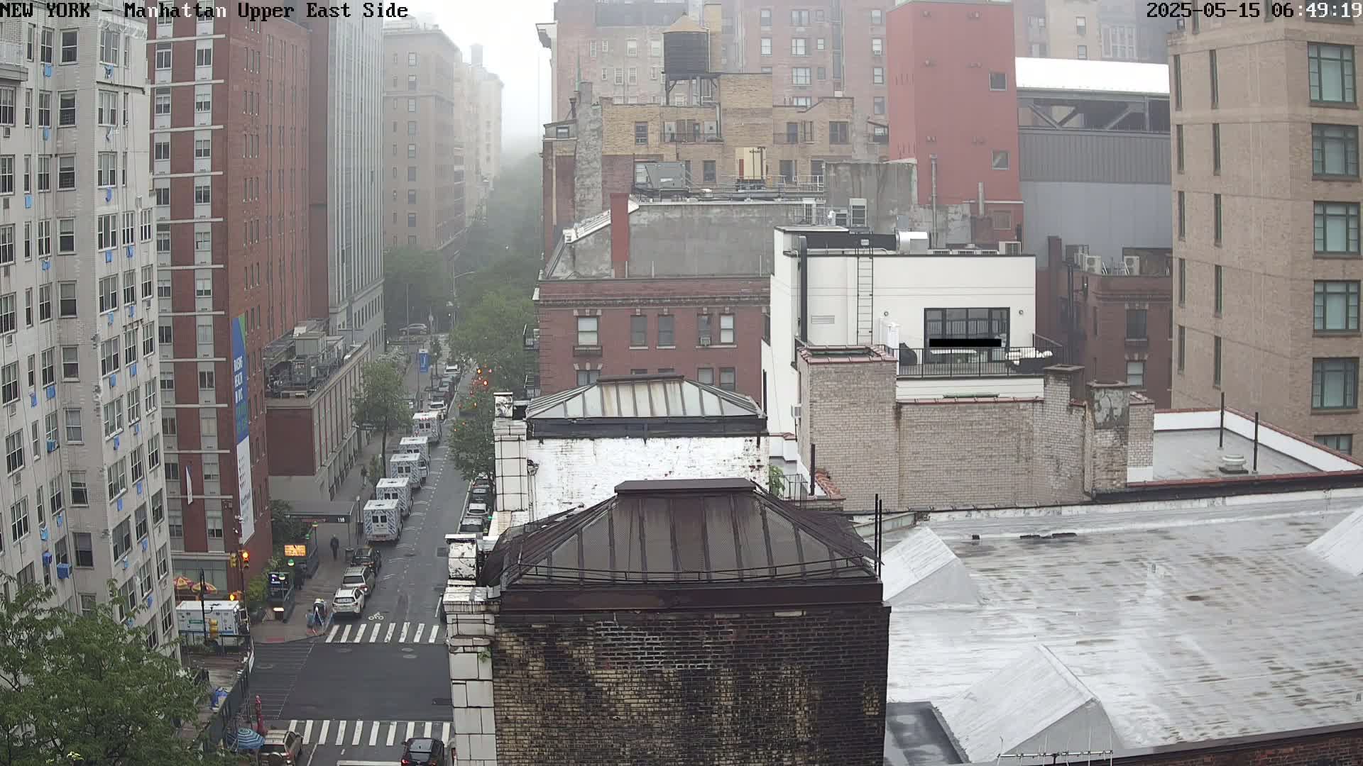 A foggy city street scene shows several multi-story buildings, a street with cars, and some trees.