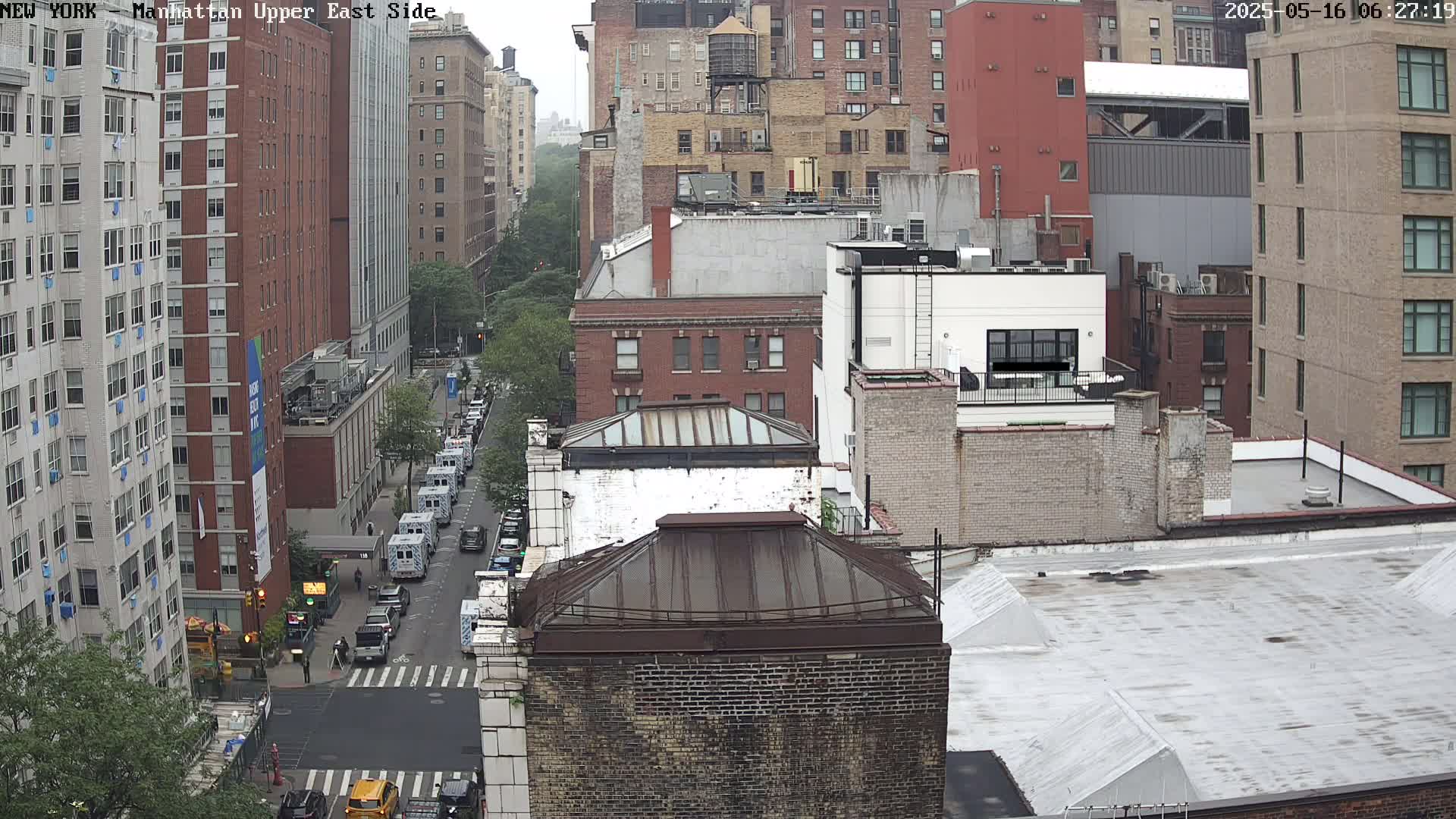 A high-angle view of a city street scene on an overcast day, showing multiple brick buildings, a street with cars and a few pedestrians, and a line of what appear to be emergency vehicles.