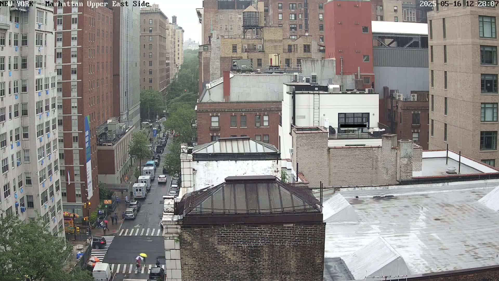 A rainy day view of a city street with several multi-story buildings, and vehicles driving down the wet road.