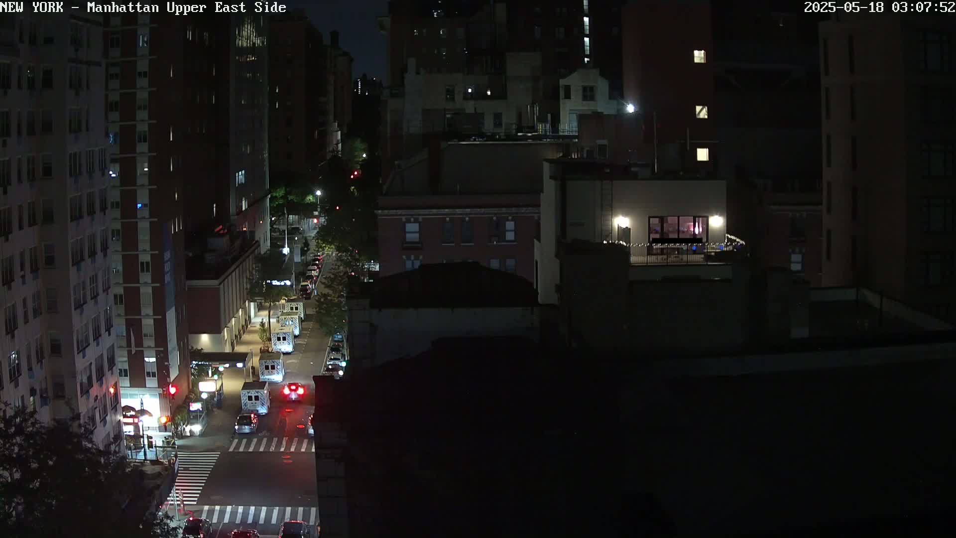 A nighttime, high-angle view of a city street shows several multi-story buildings, some vehicles on the street, and a few illuminated windows.