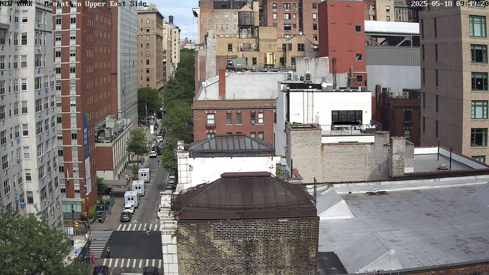 A high-angle view of city buildings on a sunny day, with a street visible below, showing some vehicles and pedestrians.