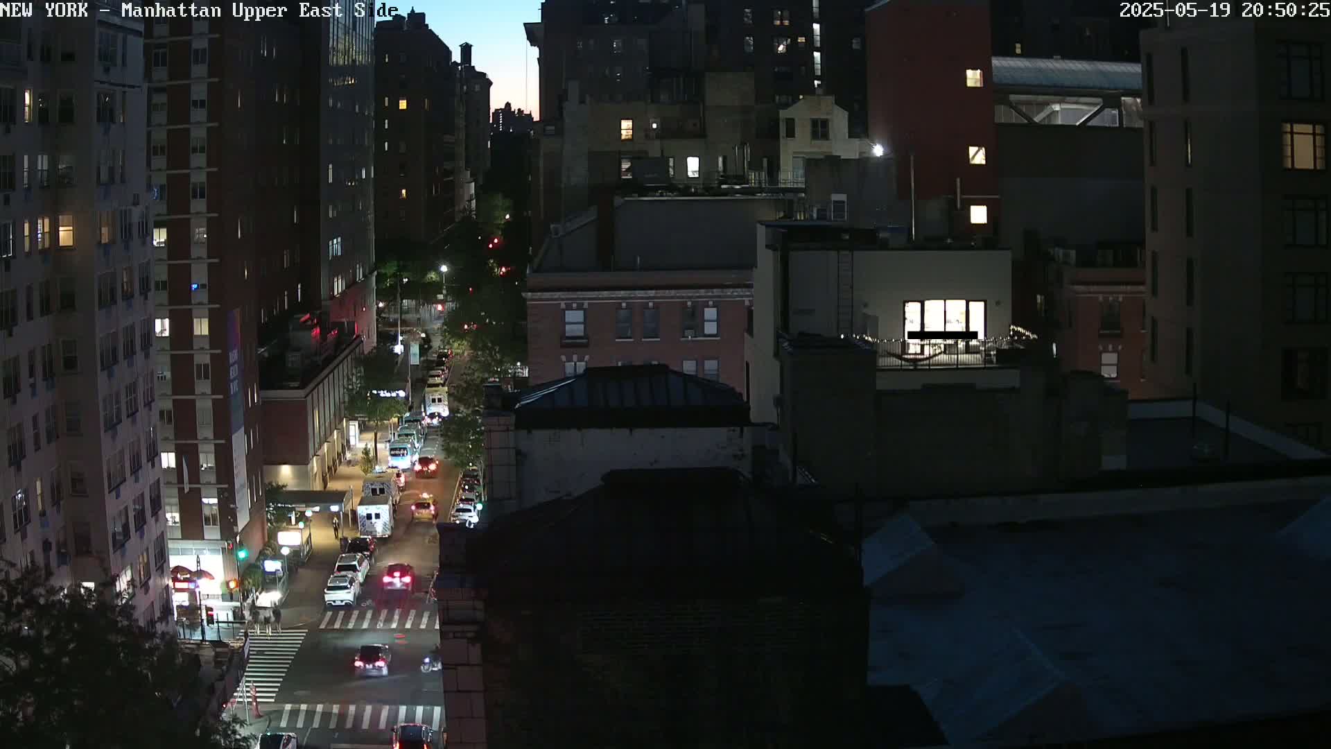 A nighttime, high-angle view of a city street with cars driving on it, surrounded by dark buildings under a twilight sky.