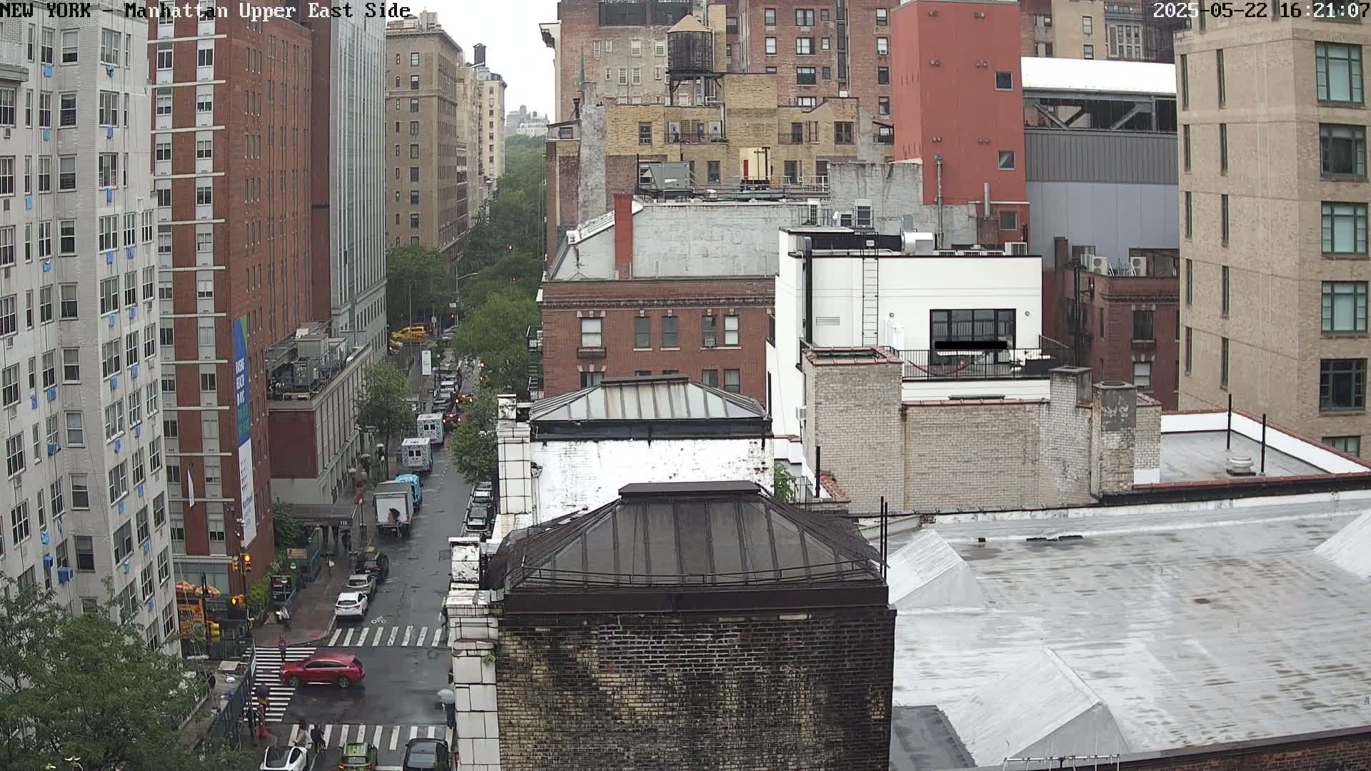 A rain-slicked city street scene shows a view of several multi-story buildings in a dense urban environment.
