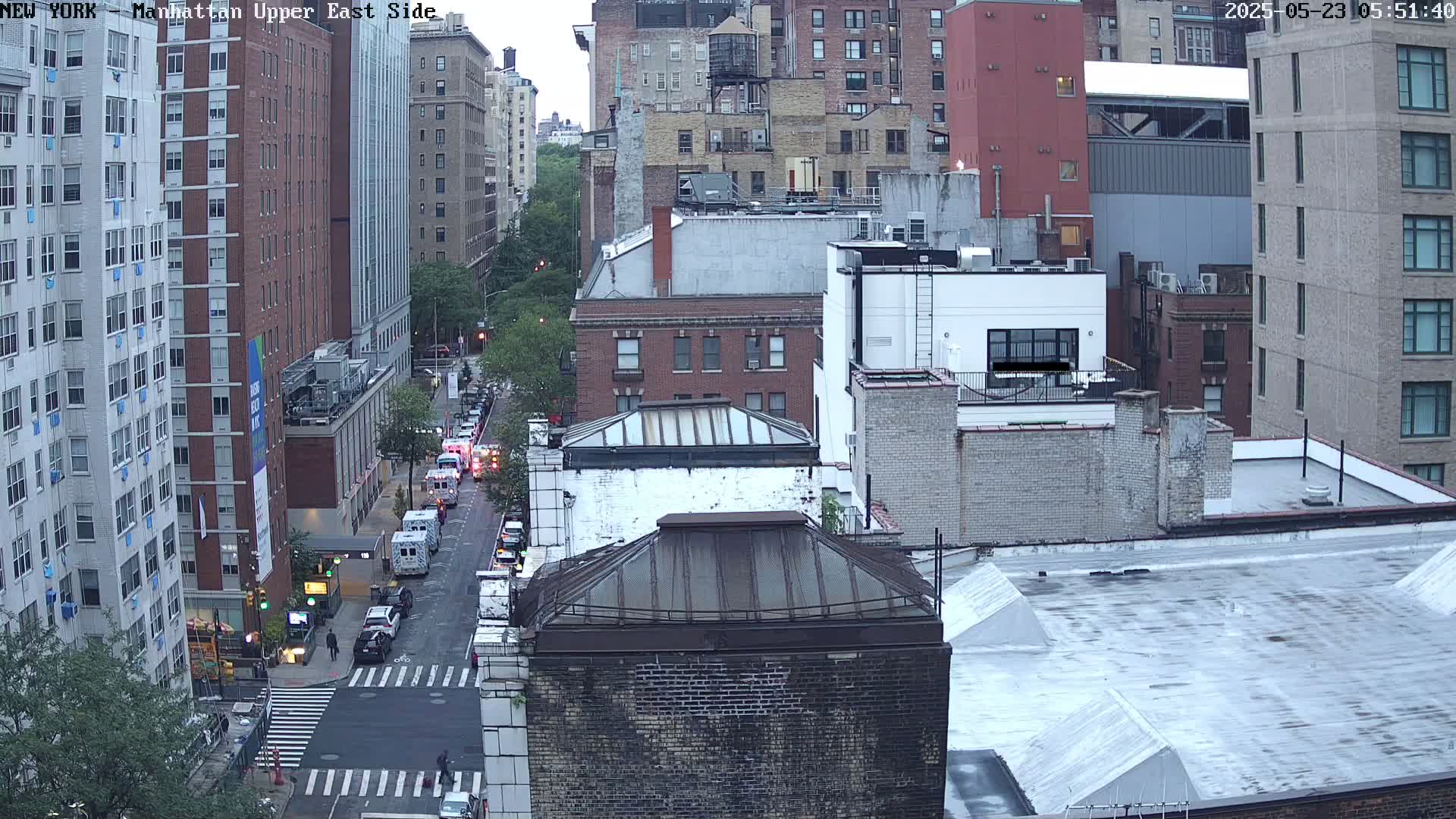 An overcast-looking city street scene with several tall buildings and a few vehicles on a street below, viewed from a high vantage point.