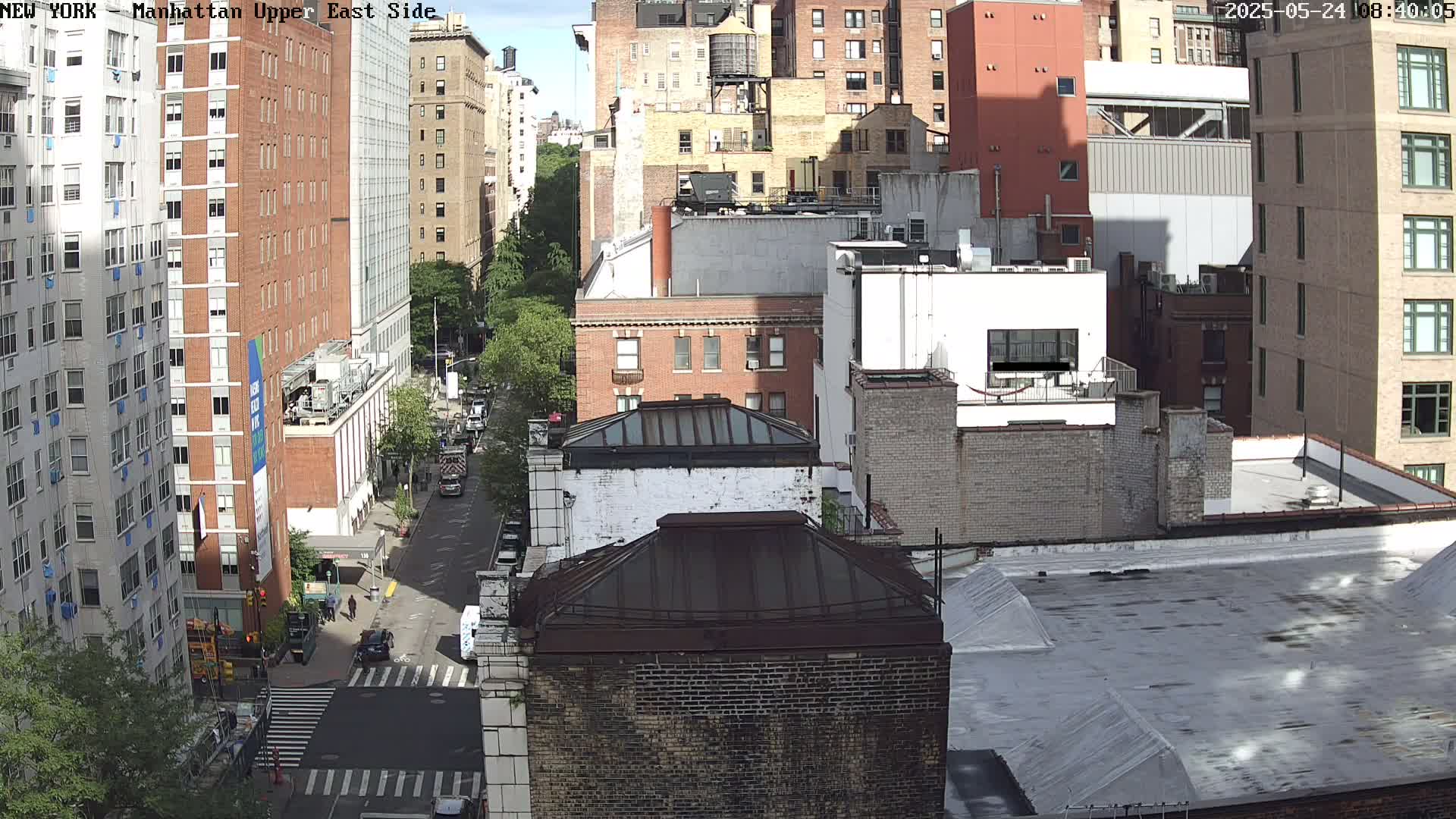 A sunny city street scene shows a high-angle view of several multi-story brick buildings and a street with sparse traffic.