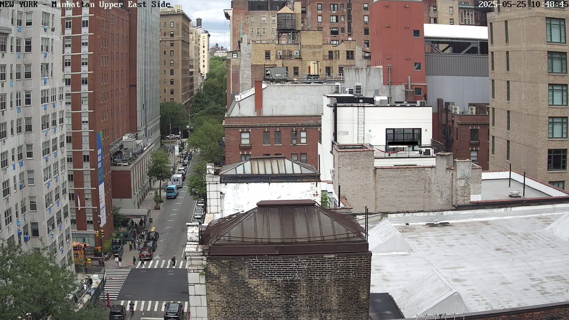 A high-angle, city view on a partly cloudy day shows numerous brick buildings, a street with cars and pedestrians, and some trees.