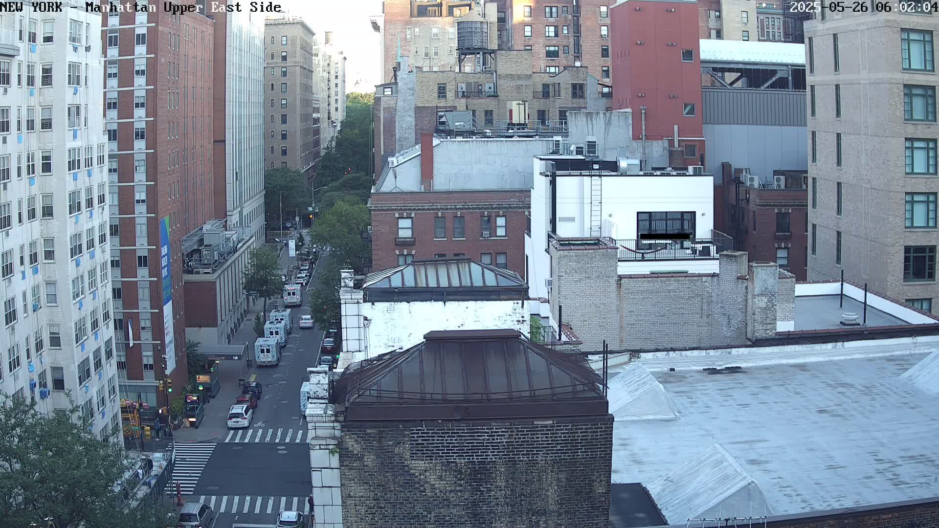 An aerial view of a city street scene shows several multi-story brick buildings under a mostly clear sky, with some cars visible on the street below.