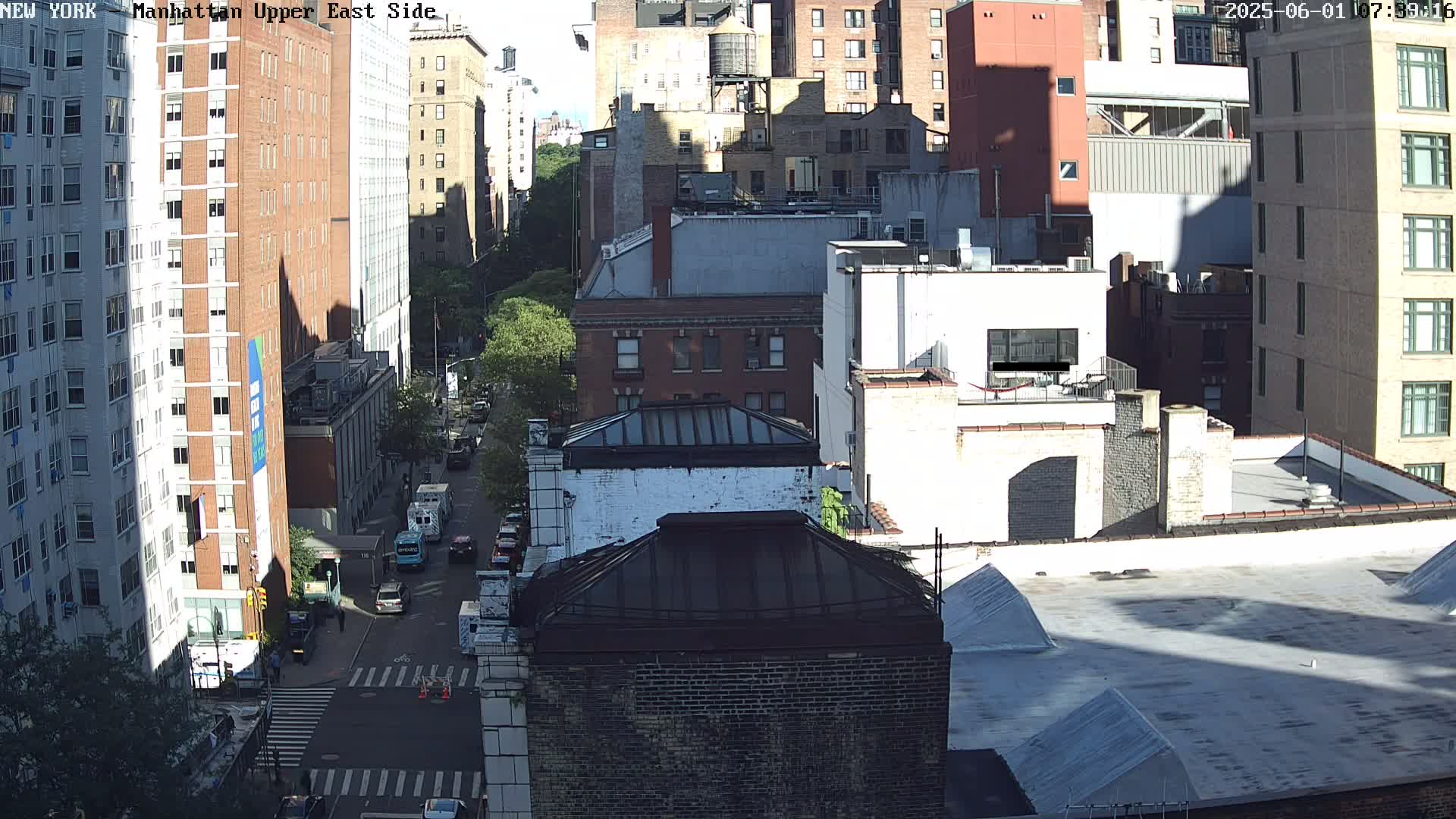 A high-angle, sunny-day view of a city street shows several multi-story buildings, with vehicles parked along the road and some pedestrians visible on a crosswalk.