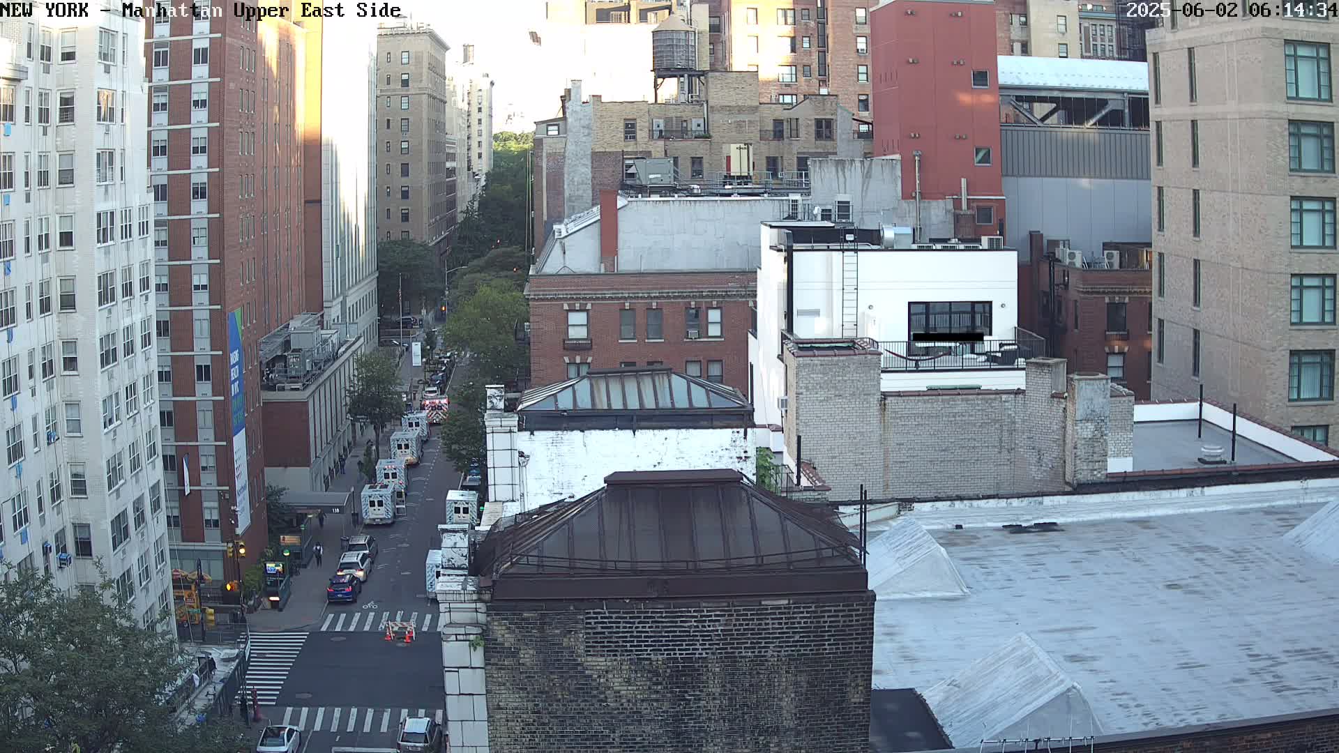 A high-angle, daytime view of a city street, showing several multi-story brick buildings and a few vehicles on the road under partly sunny skies.