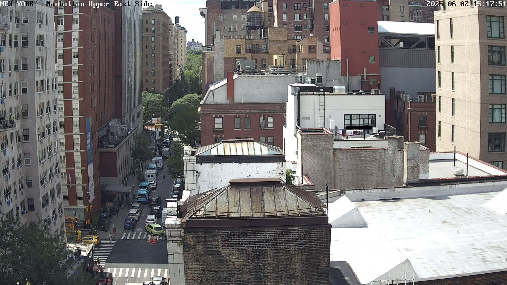 A high-angle, sunny-day view of a city street with several multi-story brick buildings, a rooftop, and parked vehicles.