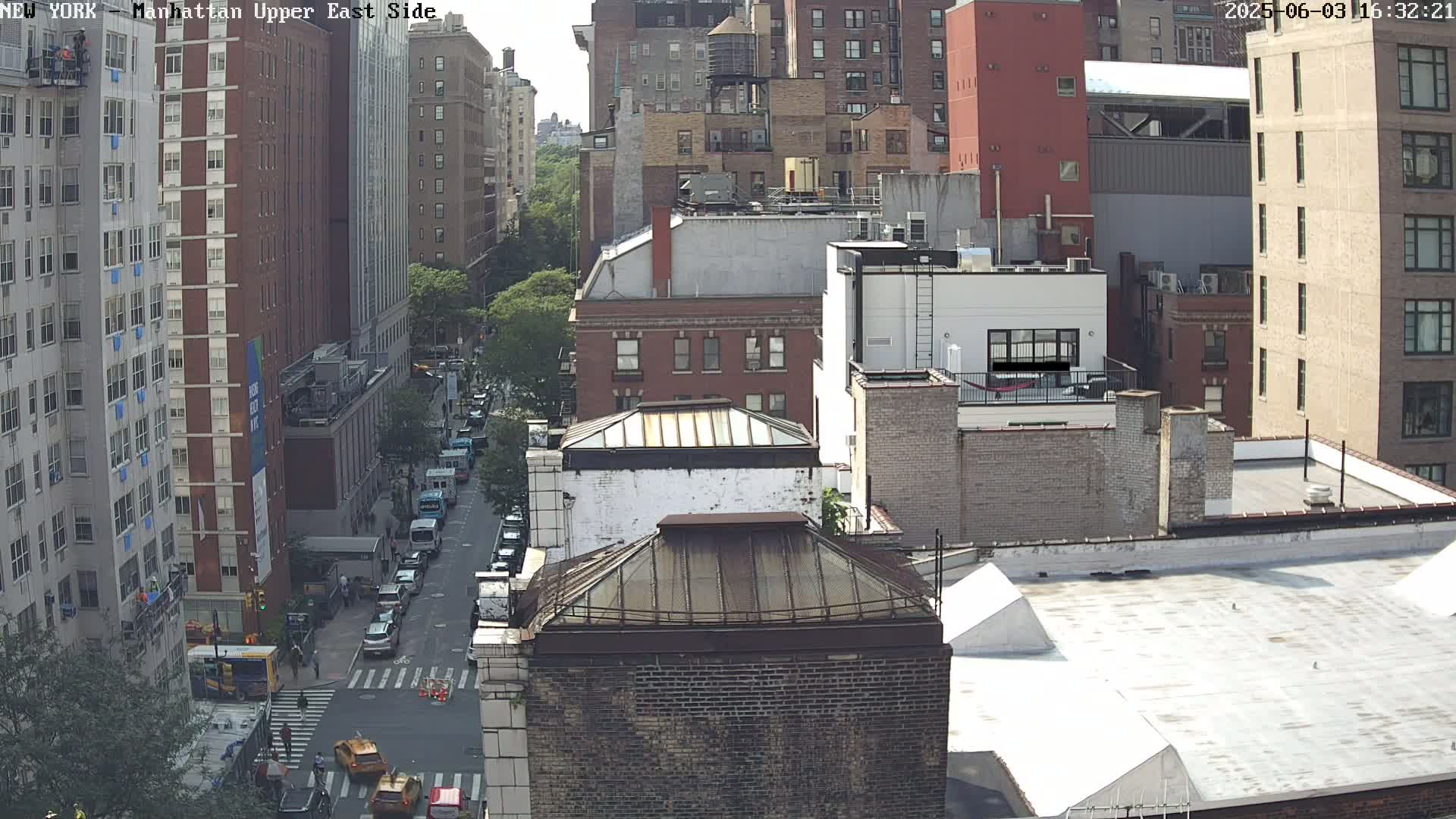 A high-angle view of a city street scene on a sunny day, showing several multi-story brick buildings, parked cars, and a few pedestrians at street level.
