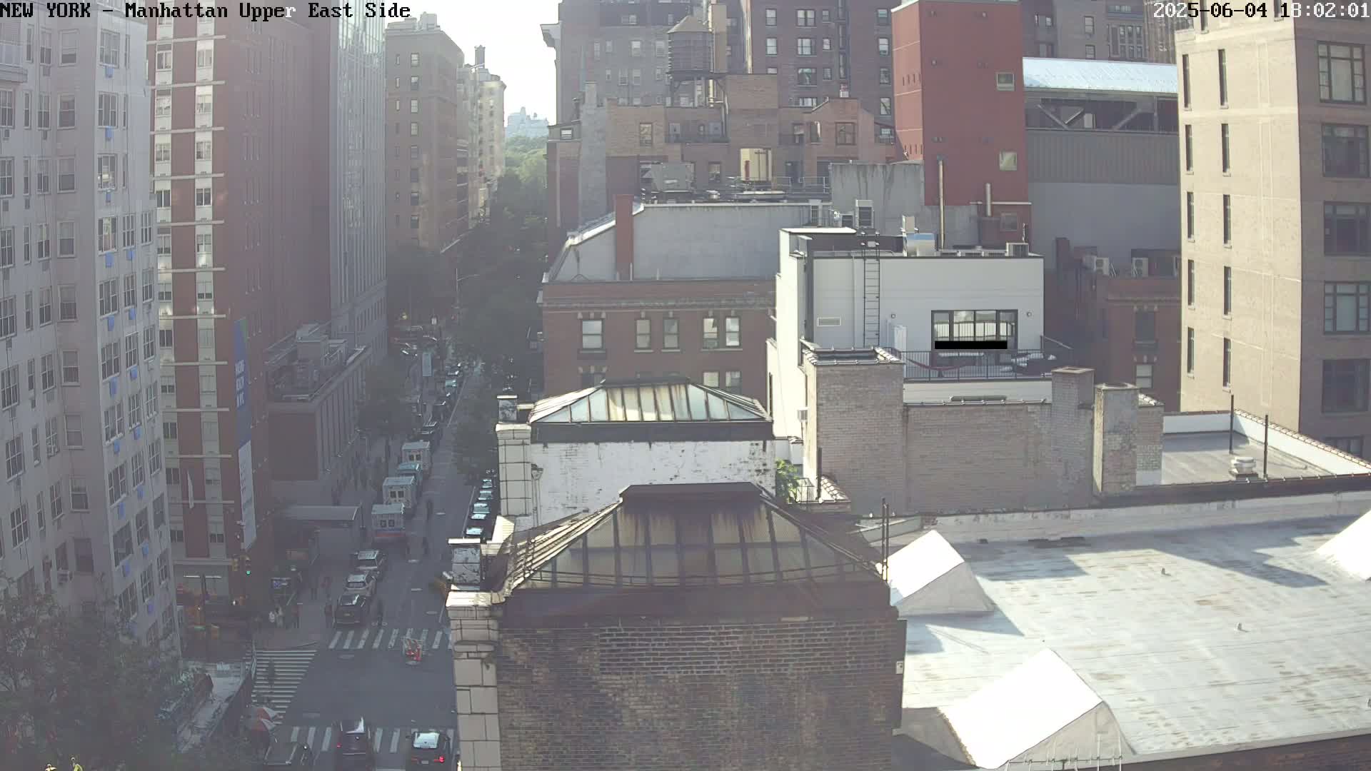 A hazy, high-angle view of city streets and buildings, showing a street with cars and pedestrians, and several multi-story buildings.