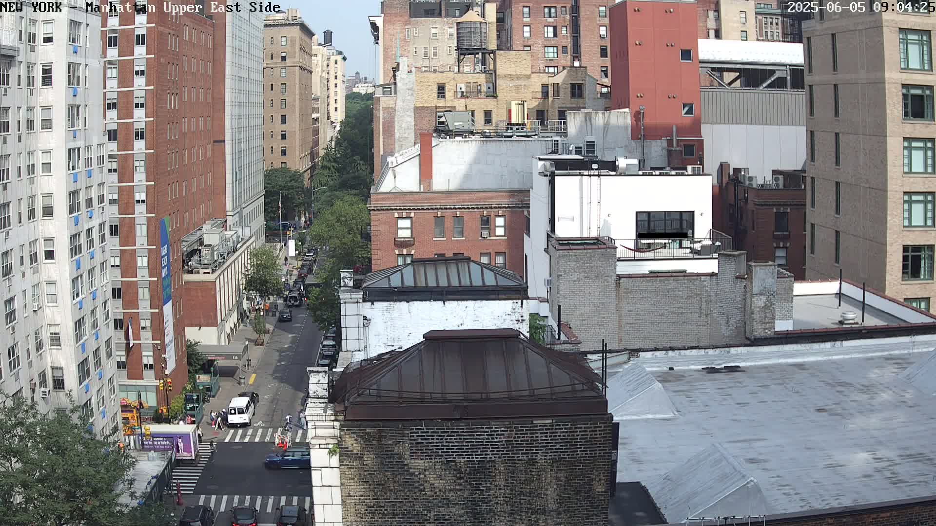 A high-angle view of a city street shows several brick buildings on a sunny day, with cars parked and driving along the road.