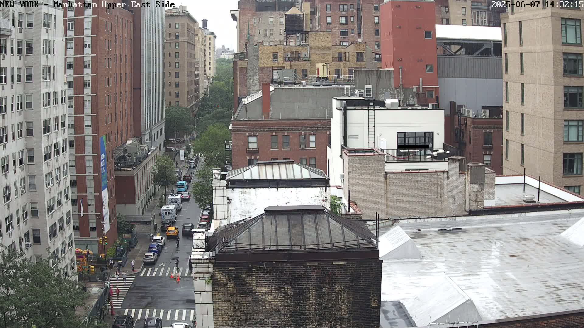 A rainy view of a city street, showing several multi-story buildings and cars driving on a wet road.