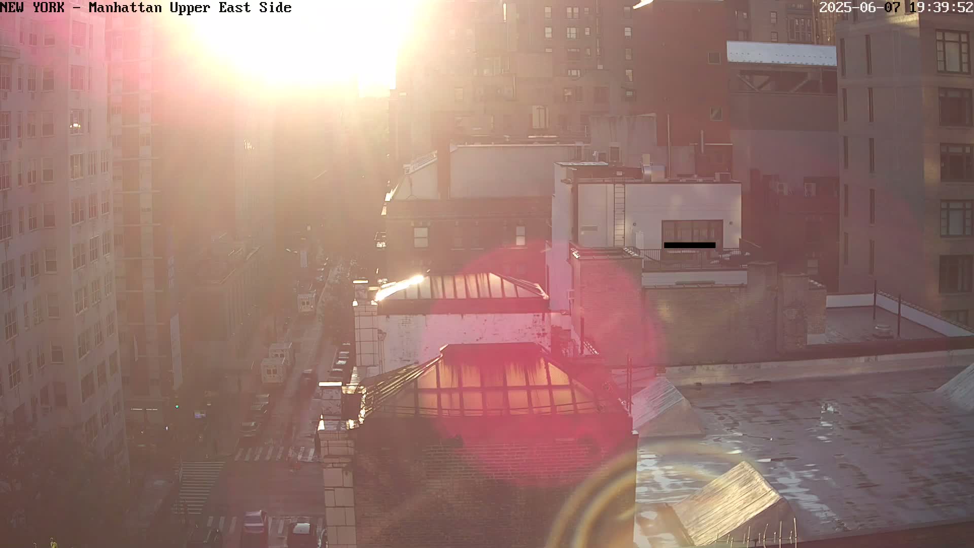 A hazy sunrise casts a warm glow over a cityscape of multi-story buildings and a street with sparse traffic.
