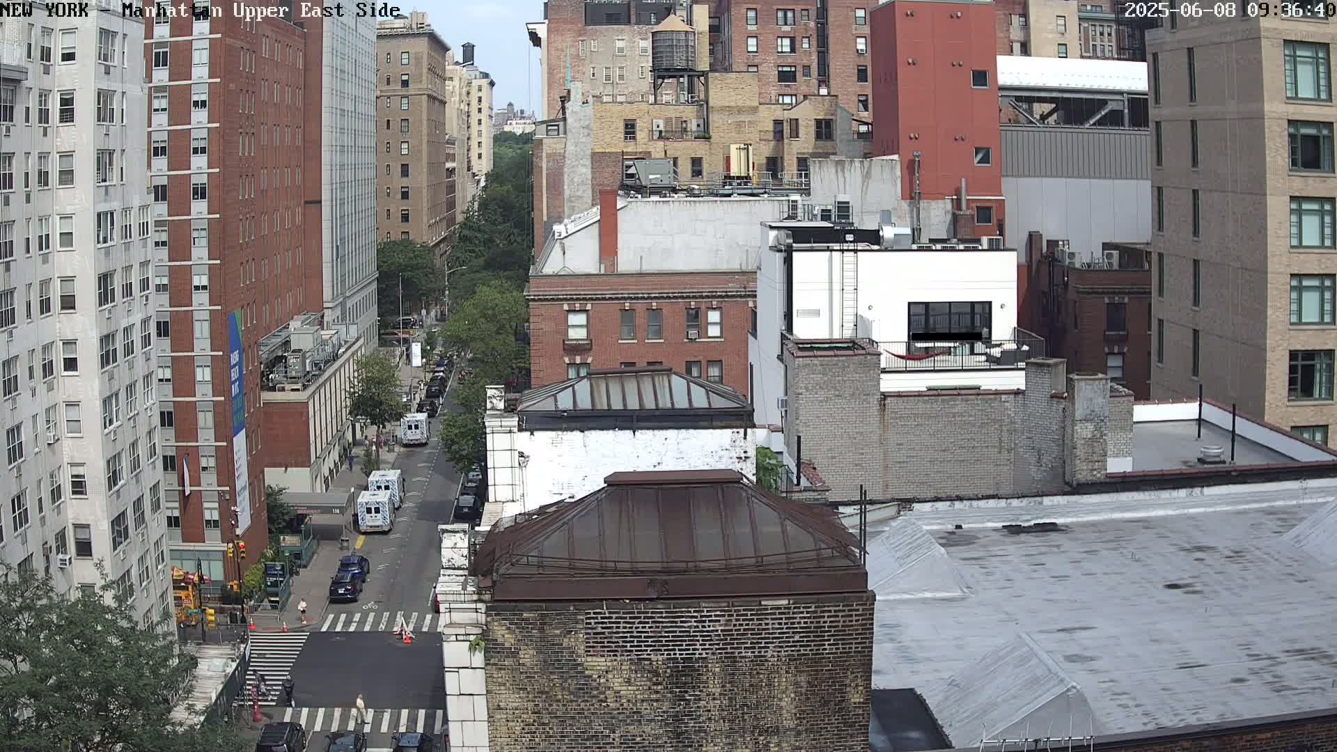 A high-angle view of a city street on a partly sunny day, showing several multi-story brick buildings, some vehicles on the street, and a few pedestrians crossing at an intersection.