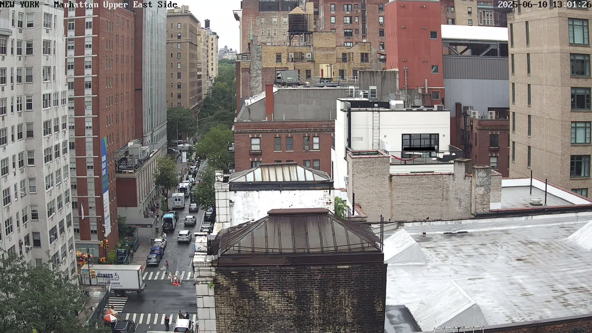A high-angle view of city streets and buildings on an overcast day, showing several cars driving along a street, and various multi-story buildings in close proximity to one another.