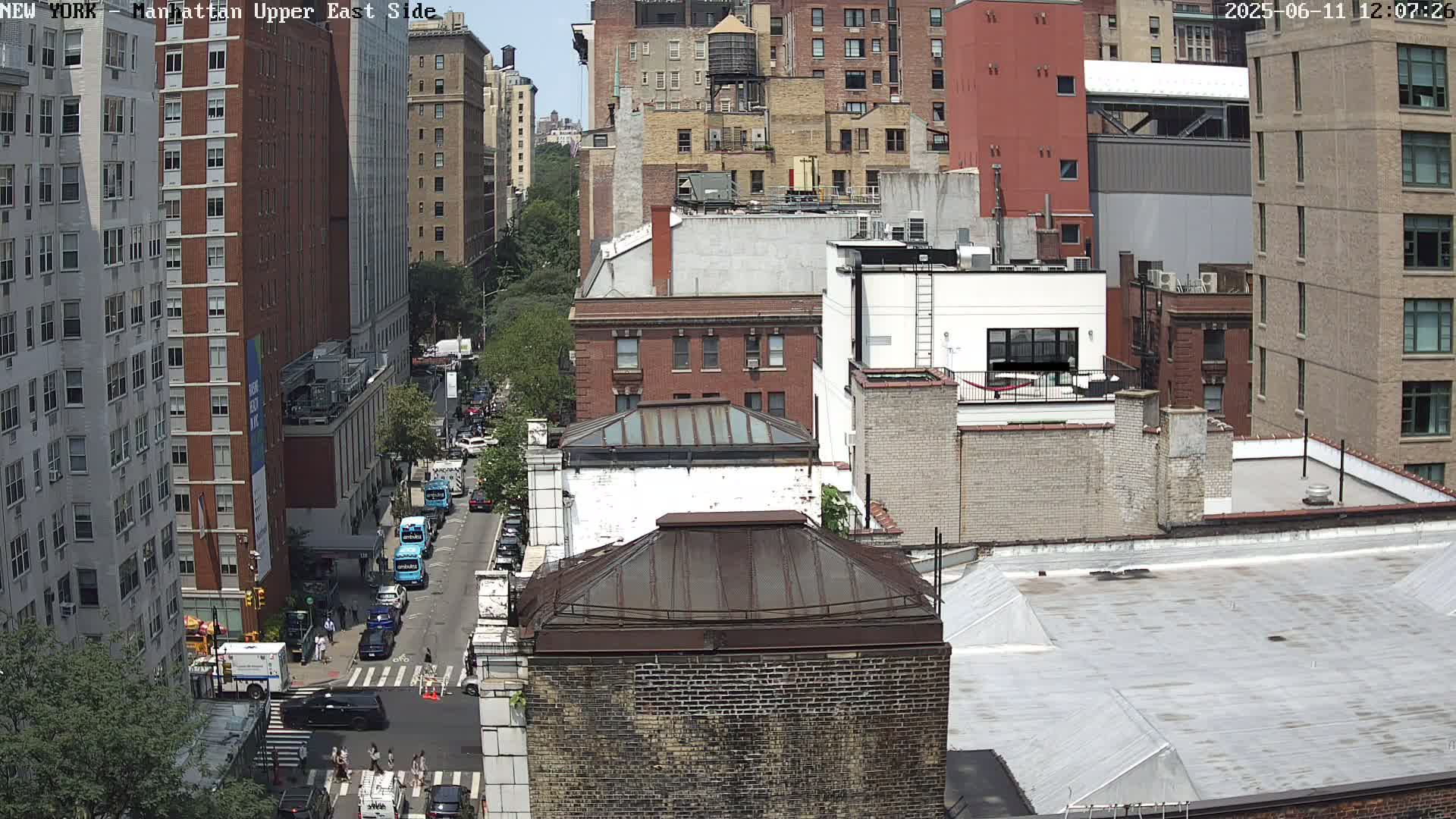 A high-angle, sunny-day view of a city street with several multi-story buildings, vehicles parked along the street, and pedestrians walking on the sidewalk.