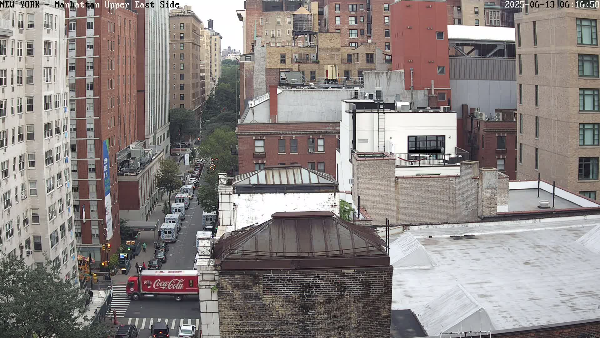 A high-angle, long shot of a city street on an overcast day, showing several multi-story brick buildings, a Coca-Cola truck, and other vehicles parked along the road.