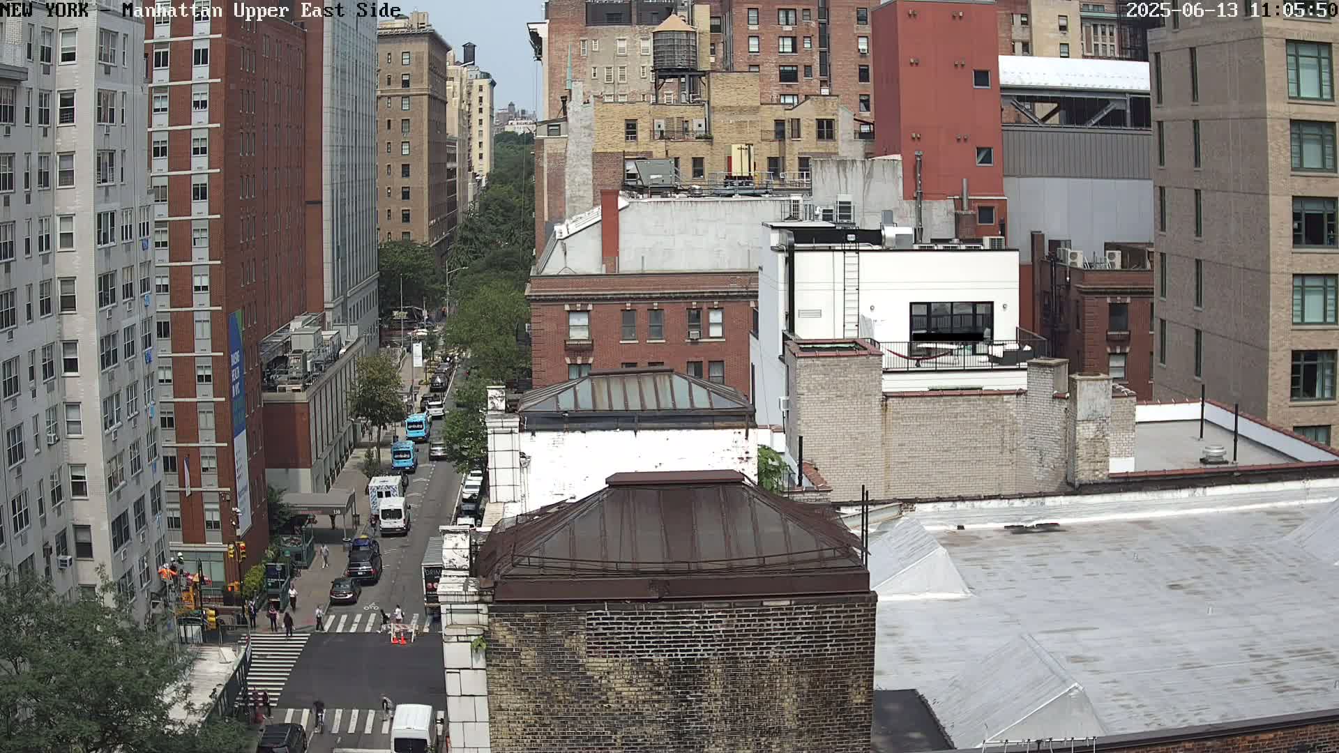 An aerial view of a city street on a partly sunny day, showing several multi-story brick buildings, cars parked along the street, and pedestrians walking on a crosswalk.