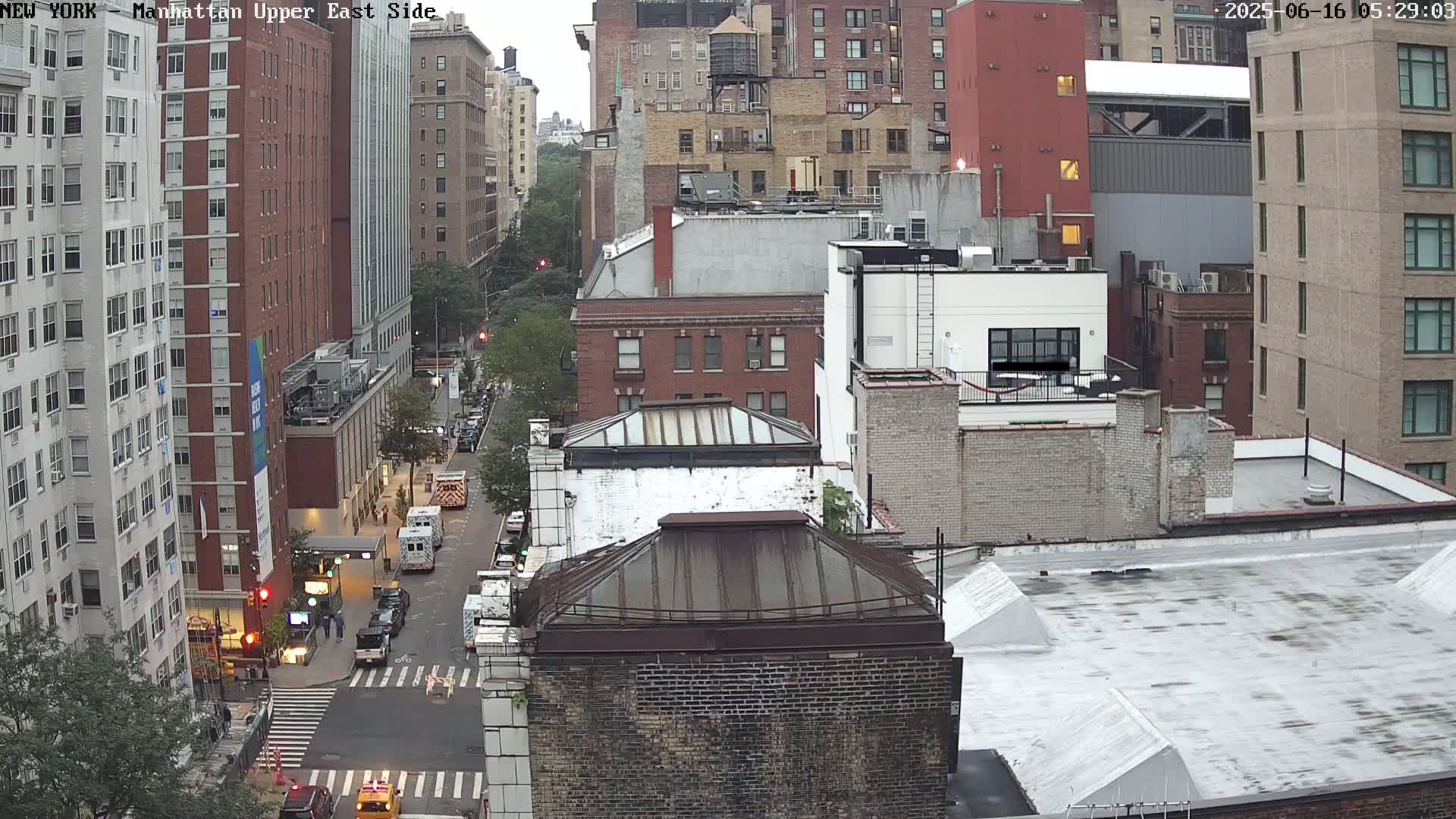 An aerial view of a city street with several multi-story buildings, overcast skies, and a few vehicles on the street.