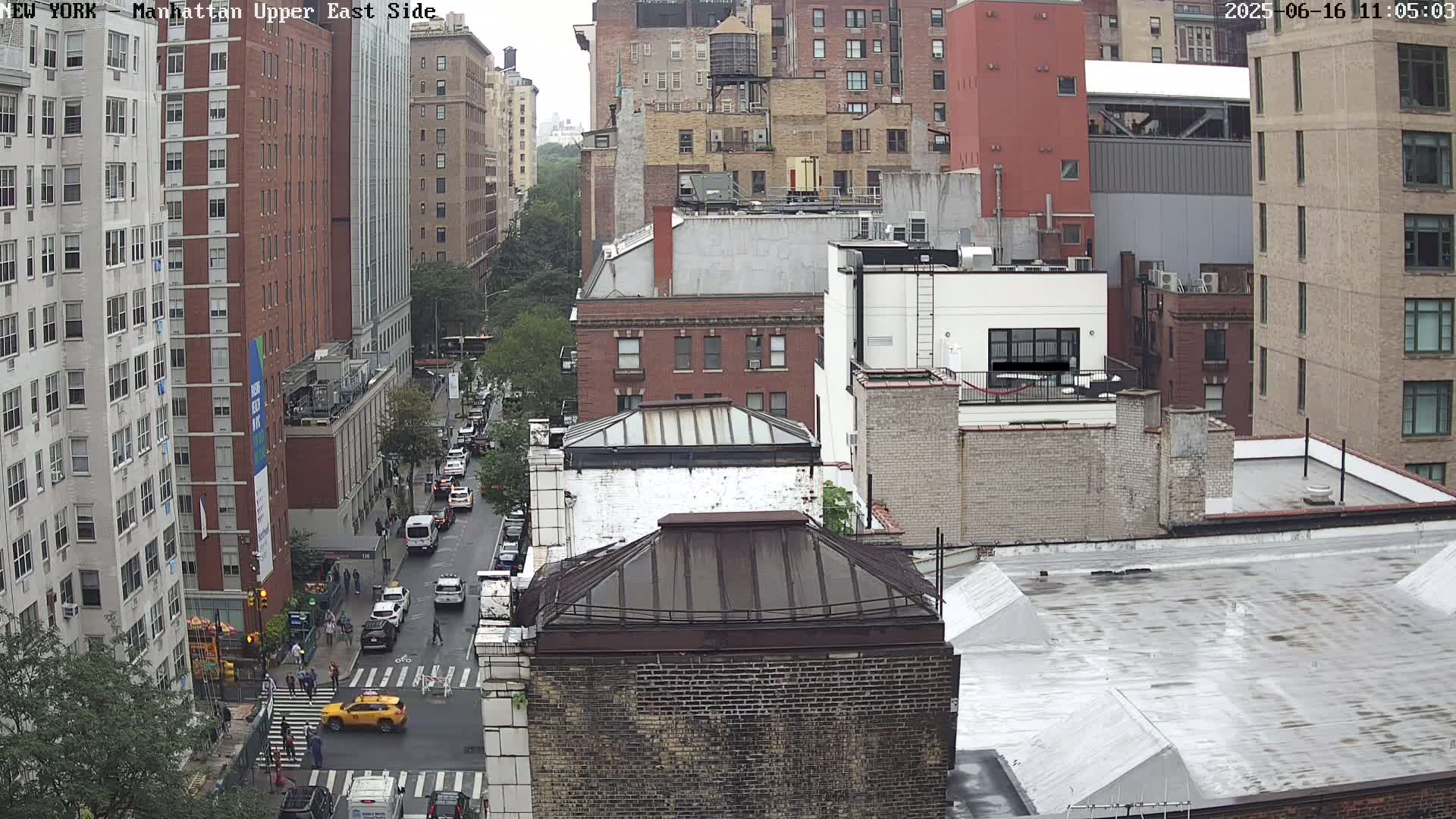 A high-angle, long shot of a city street with buildings on either side and cars parked along the road, on a cloudy day.