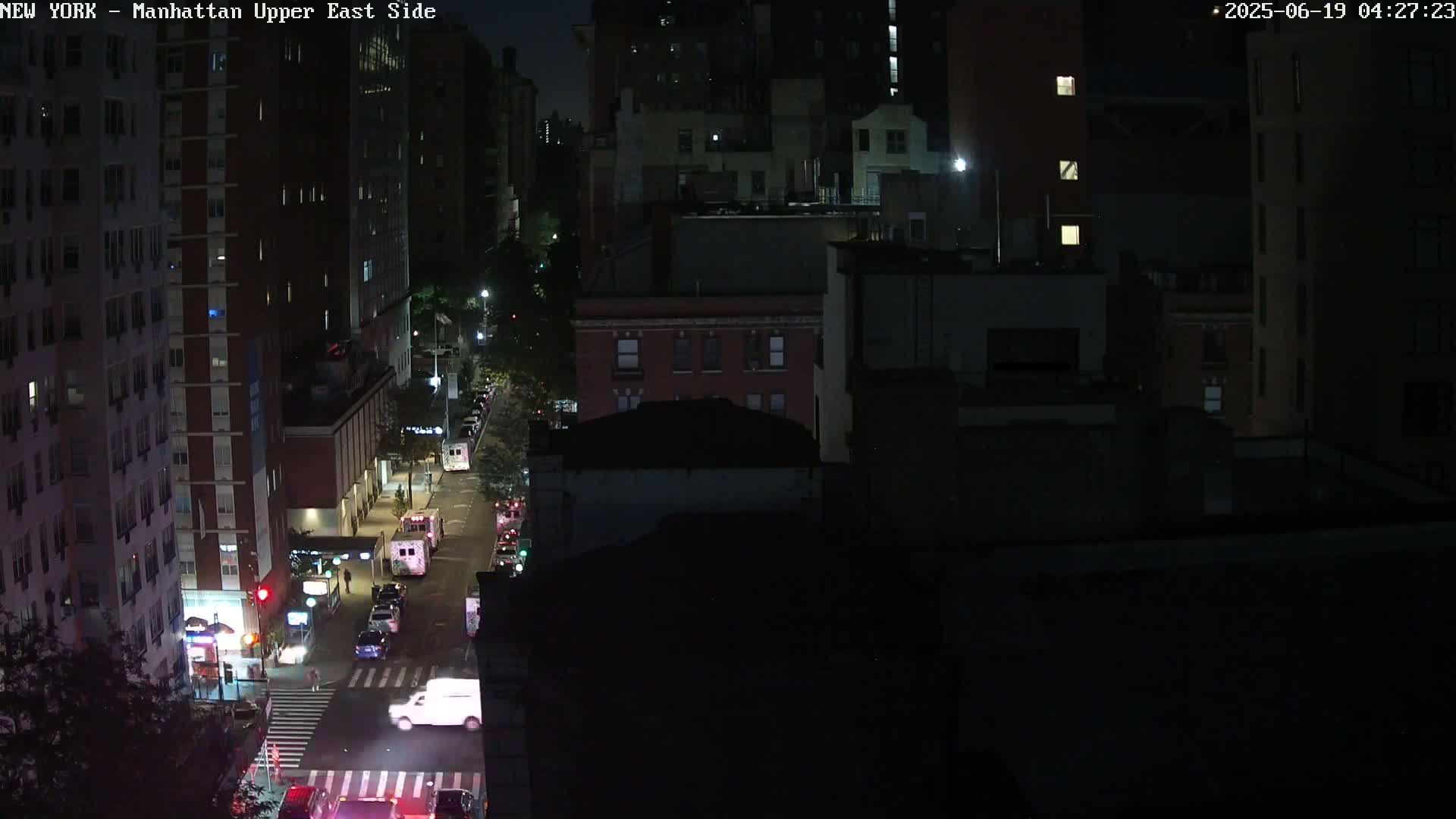 A nighttime, high-angle view of a city street with several vehicles, including what appear to be emergency vehicles, parked along the side of the road, surrounded by dark buildings.