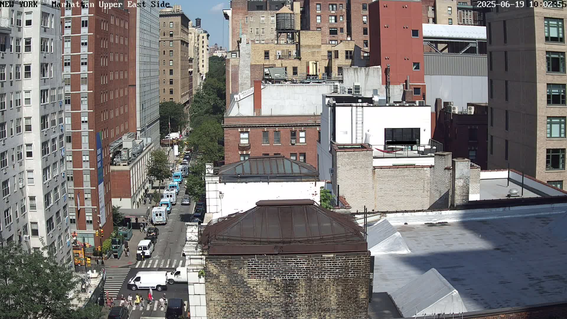 A high-angle view of a city street on a sunny day, showing several buildings, vehicles, and pedestrians at a street crossing.
