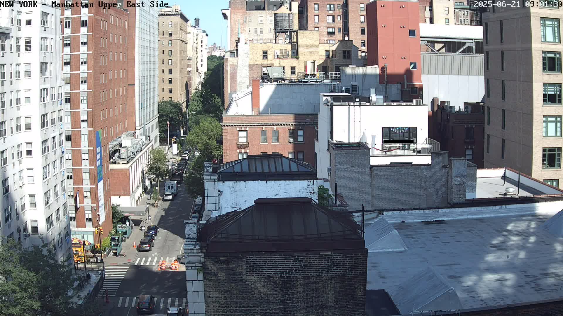 A sunny city street scene shows a view of multiple brick buildings from a high vantage point, with cars on the street below.