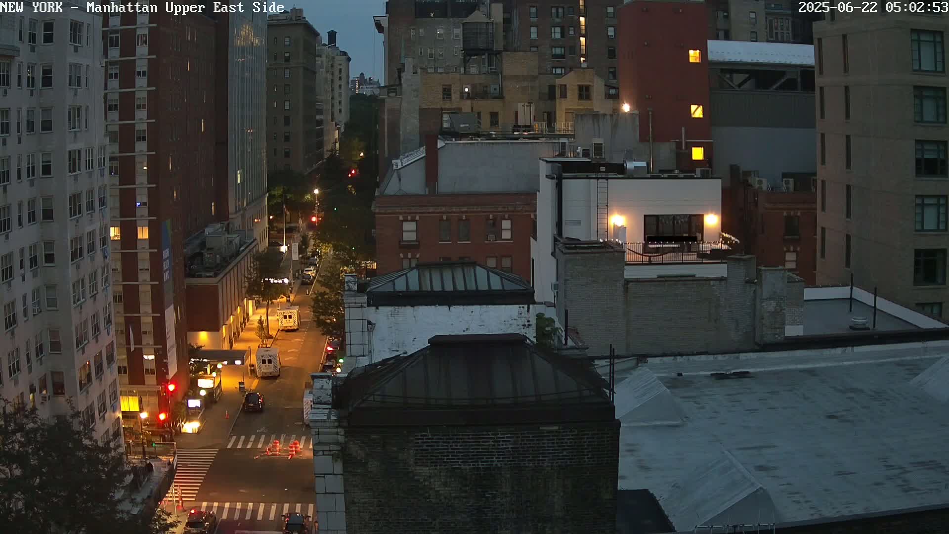 A nighttime aerial view of a city street shows several multi-story buildings, some with illuminated windows, and a few vehicles on the road.