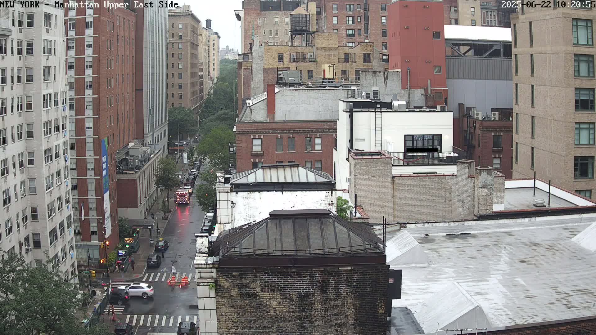 A rain-slicked city street scene shows several multi-story brick buildings under an overcast sky.