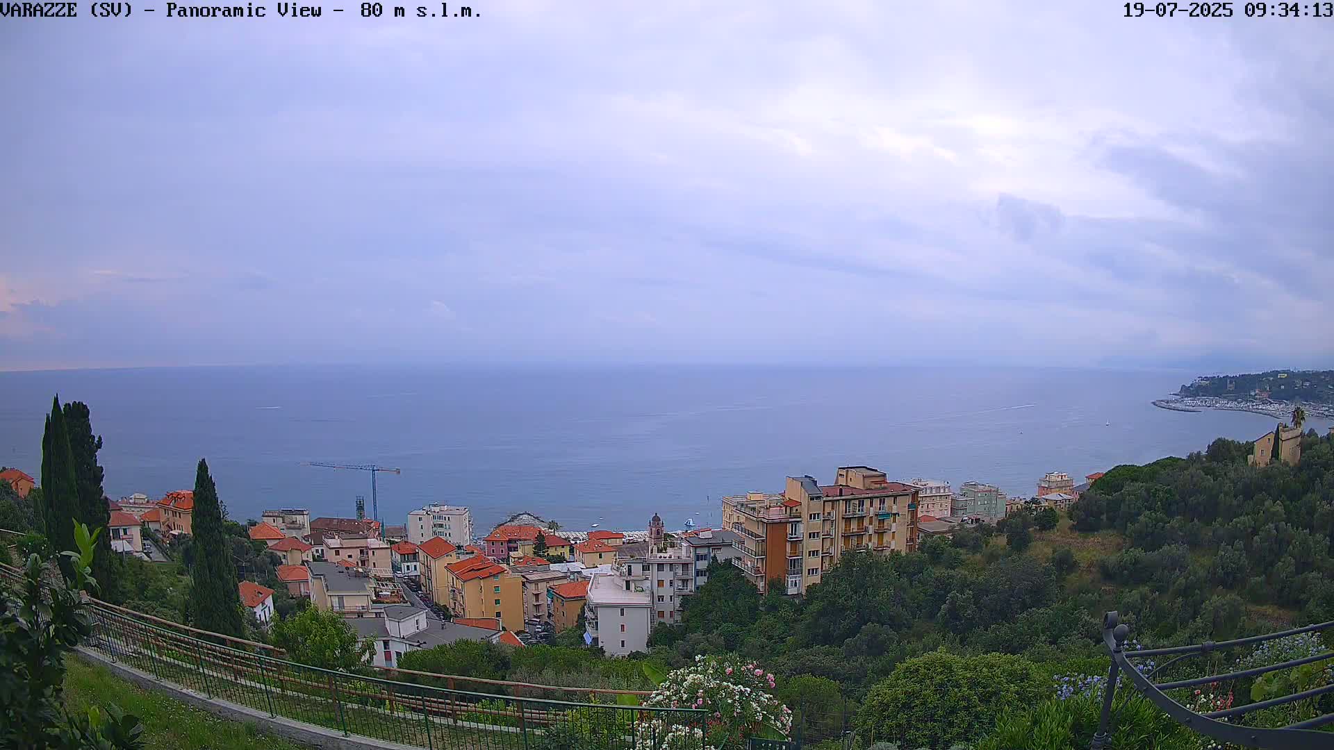 A coastal town with multi-story buildings is nestled on a hillside overlooking a calm sea under a mostly cloudy sky.