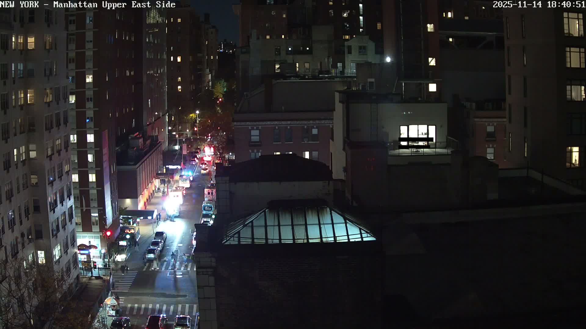 A clear night view from above shows a busy city street illuminated by streetlights and vehicle lights, including several emergency vehicles, flanked by tall buildings with many lit windows.