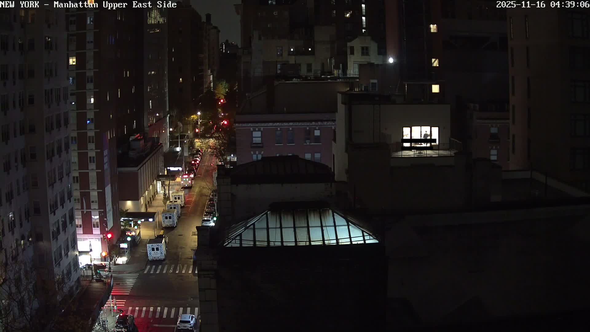 It is a clear night in a dense urban area, showcasing a street lined with numerous ambulances and cars, surrounded by multi-story buildings with some windows aglow, one revealing a person inside.