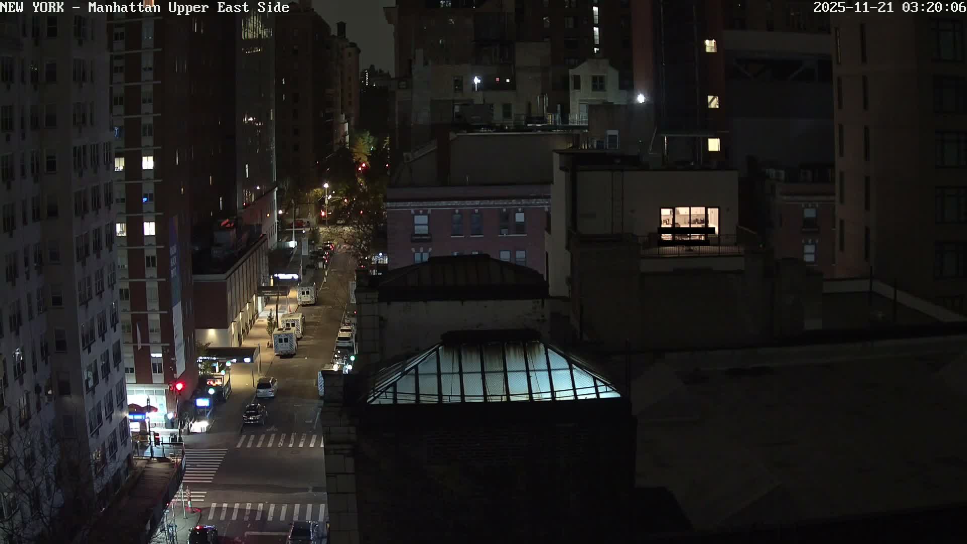 A clear night scene captures an urban street lined with multi-story buildings, featuring visible vehicles, glowing streetlights, and many illuminated windows under a dark sky.