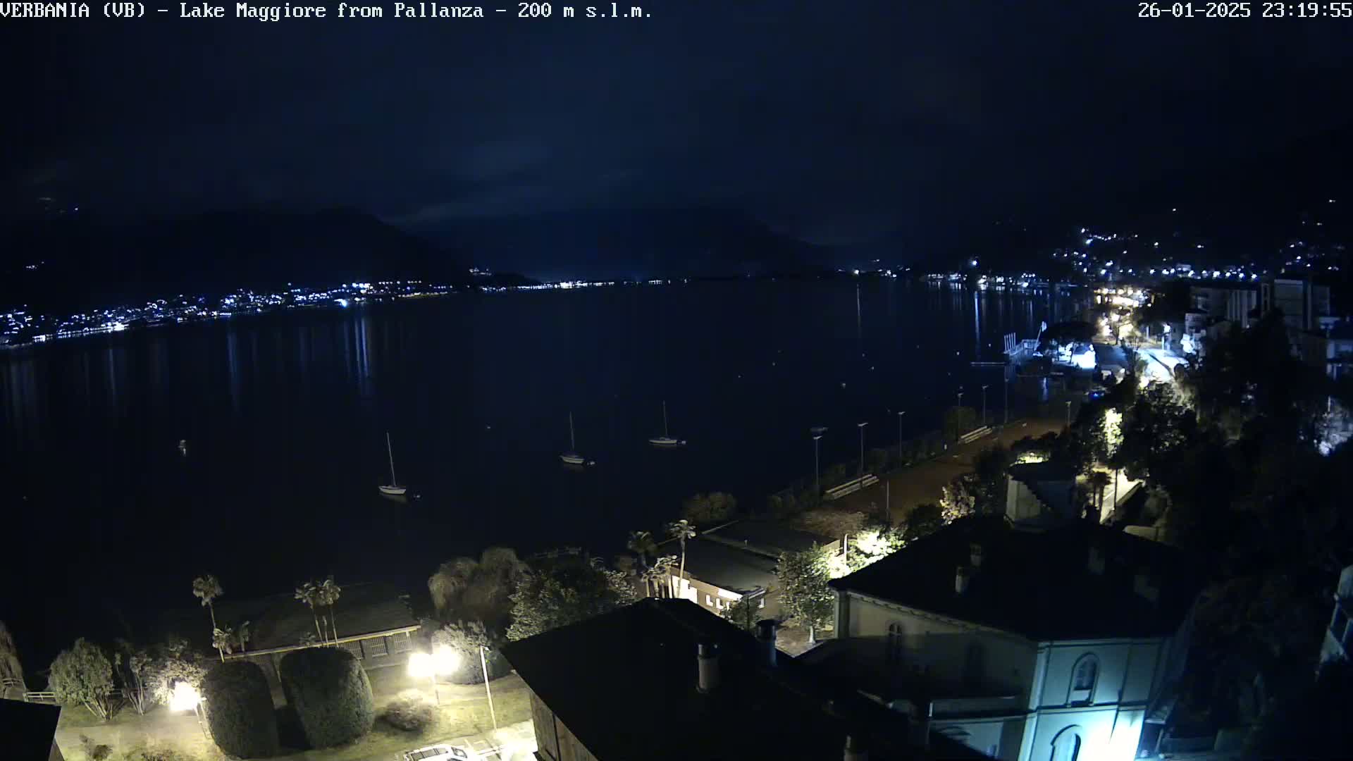 A nighttime, high-angle view shows a dark body of water with lights reflecting on its surface, boats, and a shoreline with buildings illuminated.