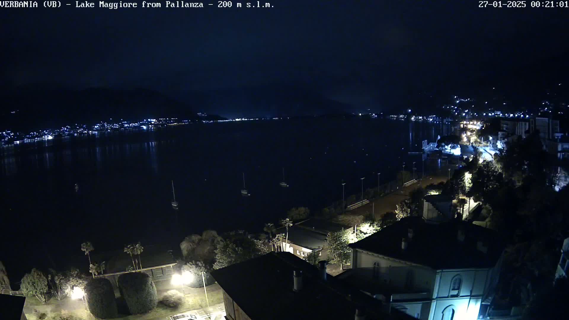 A nighttime view of Lake Maggiore from Pallanza shows a dark body of water with a few lights visible on the opposite shore, and illuminated buildings and trees in the foreground under a clear sky.