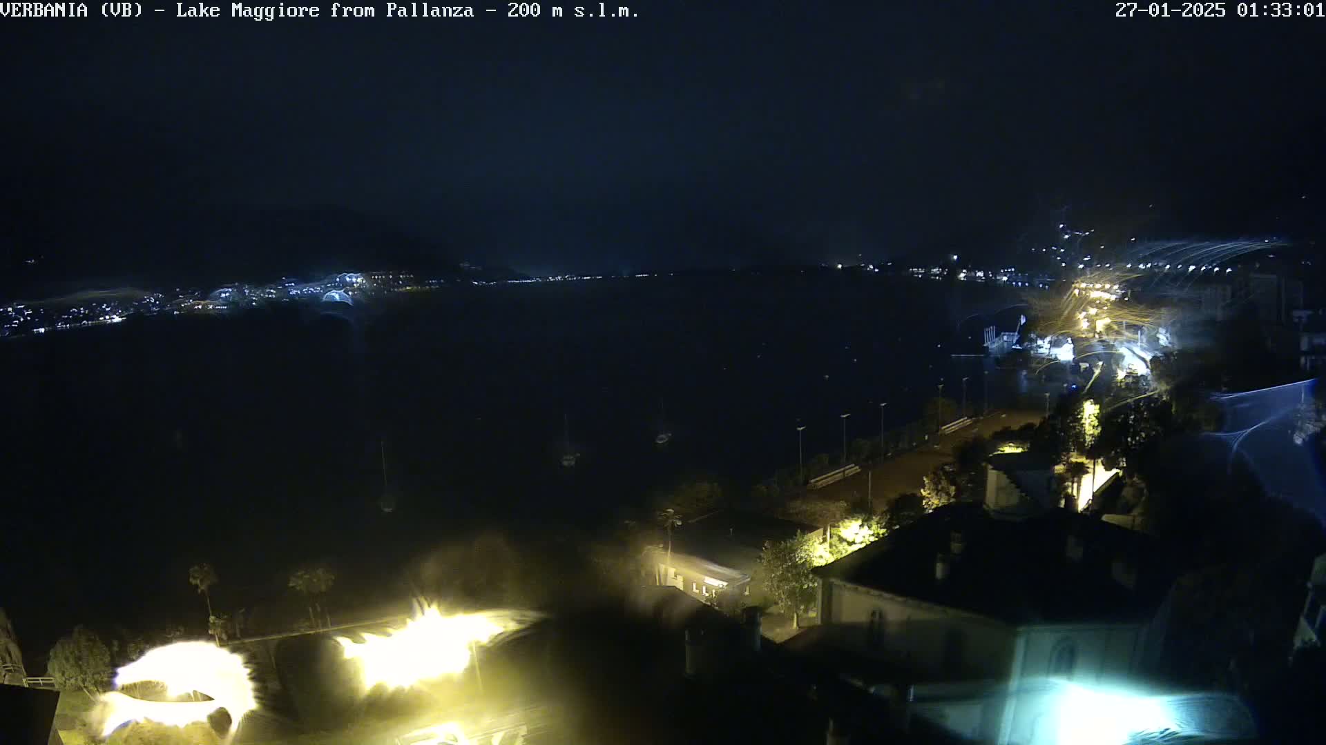 A nighttime, long shot overlooking a dark body of water with city lights in the distance and a few lights visible near the shoreline, under what appears to be windy conditions.