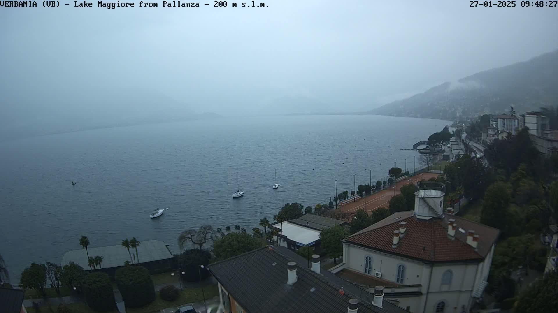 An elevated view shows a hazy, overcast day over a large lake with several small boats, a shoreline with buildings, and a tennis court.