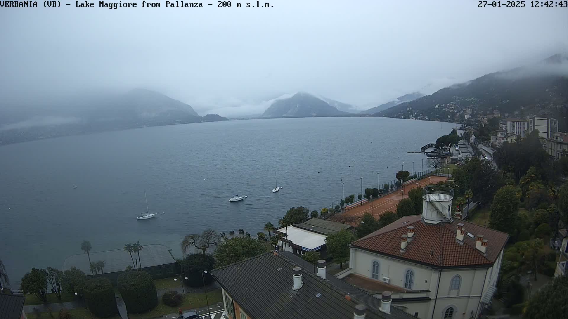An overcast day shows a view of Lake Maggiore from Pallanza, with a few boats on the water and buildings along the shoreline.
